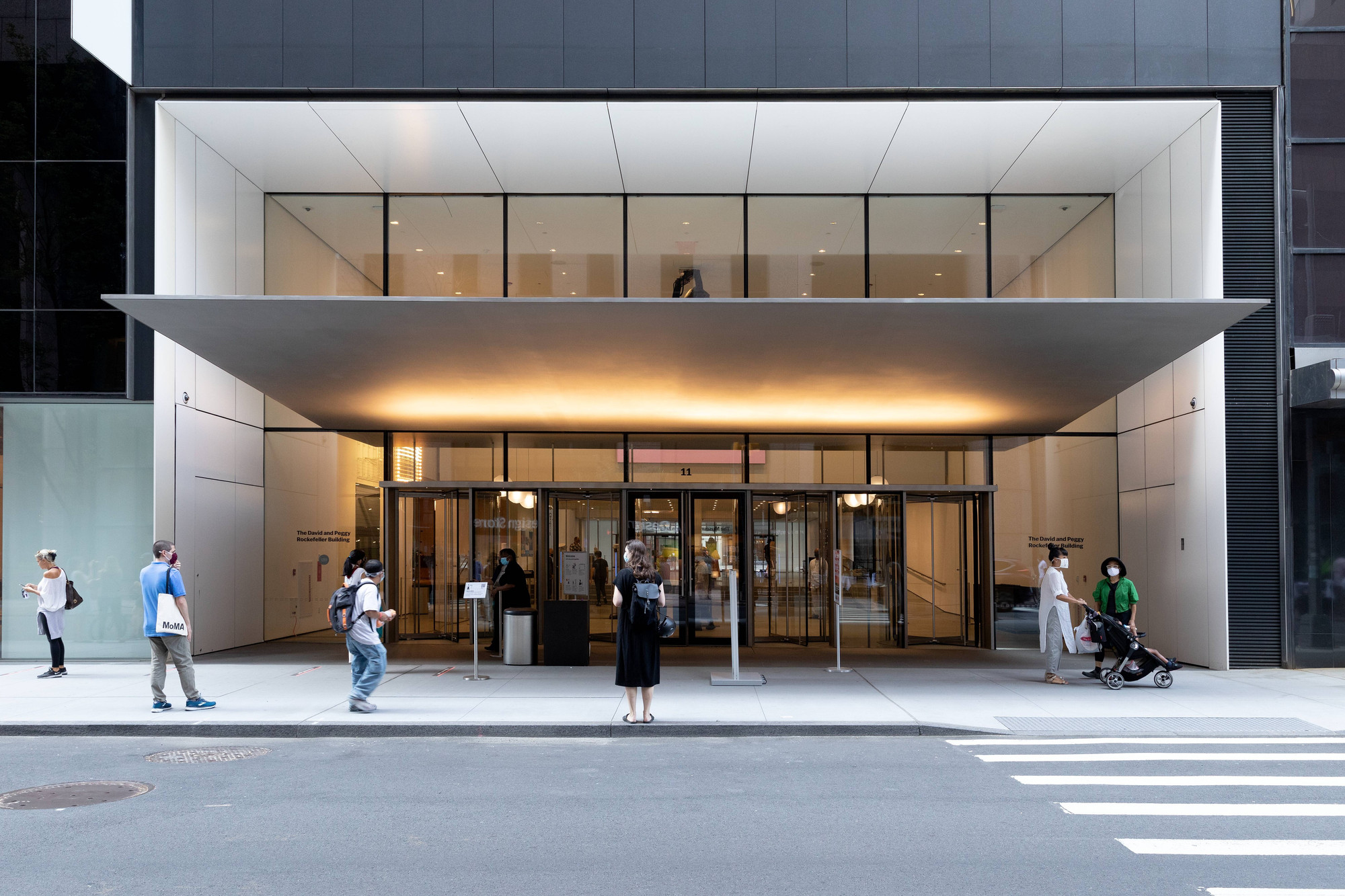 Photo: Noah KalinaImage description: MoMA’s front entrance. Glass doors with a silver rectangular overhang. A few visitors stand outside the doors. One family with a stroller exits the Museum.